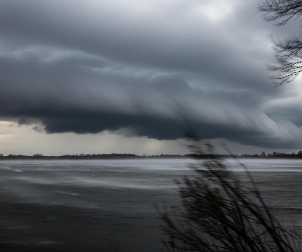 Ciemne, burzowe chmury dominują niebo, z wyraźną, poszarpaną strukturą wskazującą na ruch i silny wiatr. Poniżej, na horyzoncie, rozciąga się pas ciemnych drzew lub krzewów, a nad wodą widać warstwę mgły lub zamglenia. Powierzchnia wody jest wzburzona, z rozmytymi białymi smugami, które sugerują intensywne fale lub pęd wiatru. Po prawej stronie kadru widoczne są rozmyte, nagie gałęzie drzew, które dodatkowo podkreślają dynamiczny charakter sceny.