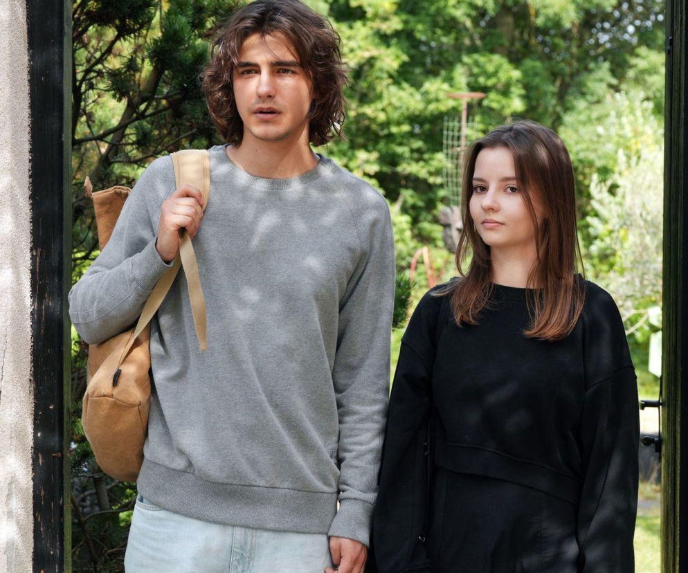 Romeo (Jacek Tyszkiewicz) w szarej bluzie z plecakiem i Ewa (Gabriela Ziembicka) w czarnej bluzce, patrzą przed siebie z zamyśleniem. Młodzi bohaterowie serialu Barwy szczęścia po ślubie mierzą się z niechęcią rodzin. Więcej o ich losach na stronie Super Seriale.