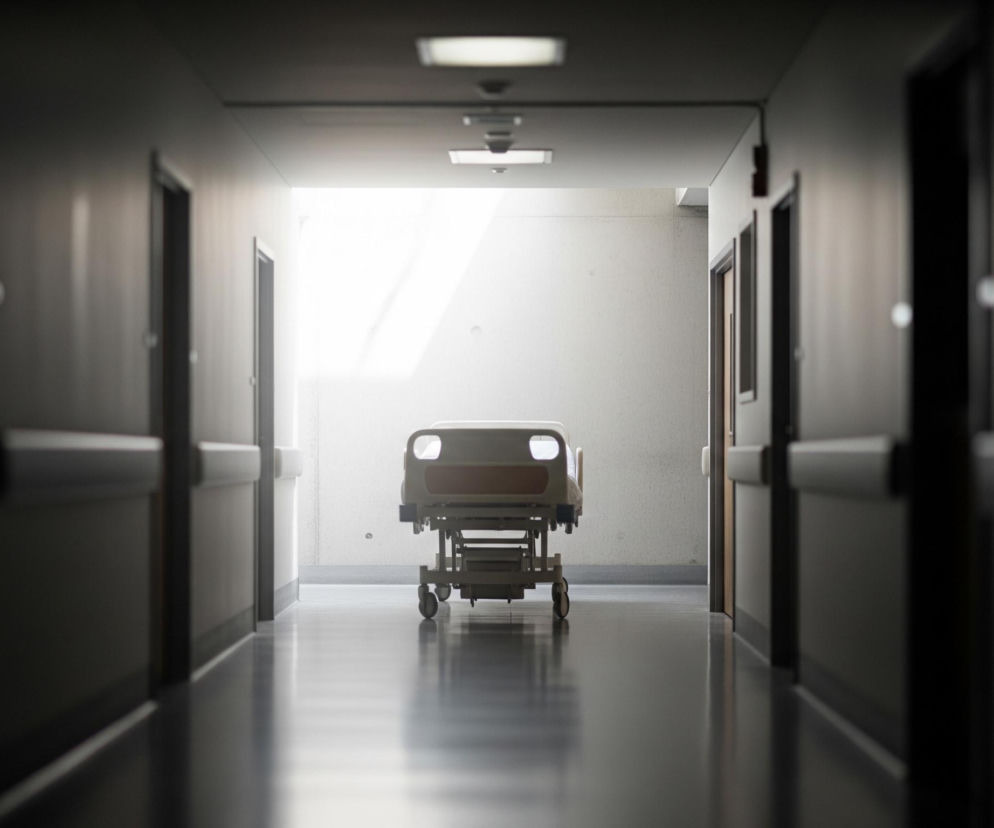 Na wprost, na końcu długiego, słabo oświetlonego korytarza, stoi puste łóżko szpitalne na kółkach. Jasne, białe światło wdziera się zza łóżka, tworząc kontrast z ciemniejszymi ścianami i podłogą korytarza. Po obu stronach korytarza widoczne są zamknięte drzwi, a na ścianach biegną jasnoszare, poziome poręcze. Sufit korytarza wyposażony jest w prostokątne oprawy oświetleniowe, które rzucają światło na powierzchnię.