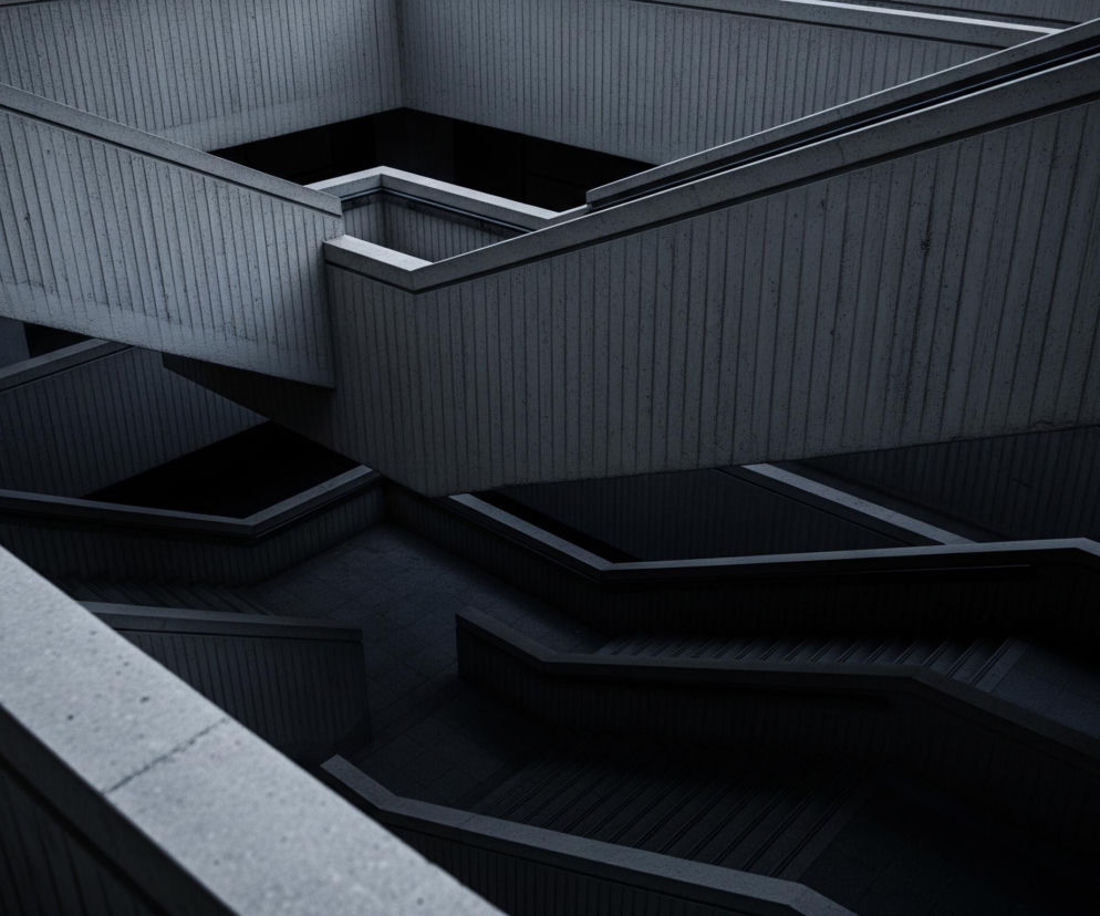 Na zdjęciu widoczna jest struktura architektoniczna złożona z betonowych elementów, przedstawiona z perspektywy z góry, w tonacji szarości z akcentami niebieskiego. Liczne, poziome i ukośne linie tworzą dynamiczny labirynt schodów i balustrad, które częściowo zanurzają się w cieniu, tworząc głębię i kontrast. Faktura betonu jest chropowata, z widocznymi pionowymi rowkami na niektórych powierzchniach. Kolorystyka jest ciemna, dominują odcienie grafitu i ciemnego błękitu, z jaśniejszymi fragmentami betonu pod wpływem światła.