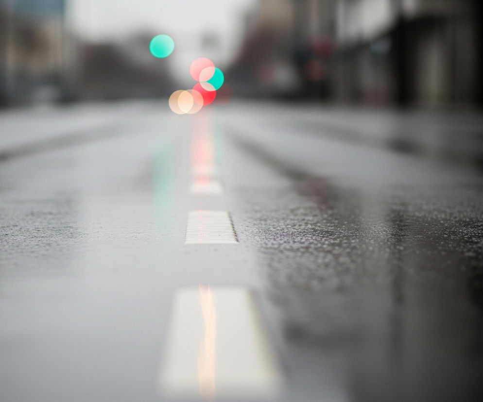 Mokra nawierzchnia ulicy dominującej szarością ciągnie się przez całe ujęcie. Na środku widoczna jest przerywana biała linia, której poszczególne segmenty rozmazują się w odbiciach kolorowych świateł w oddali. Tło pozostaje niewyraźne, ukazując rozmyte kształty budynków po obu stronach drogi oraz liczne, okrągłe, rozproszone światła w kolorach turkusowym, czerwonym i pomarańczowym.
