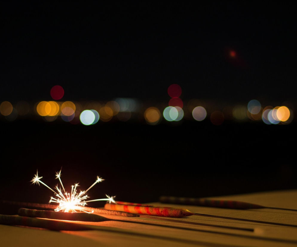 Na pierwszym planie, w dolnej części kadru, leży zapalona zimna ognia, z której iskry strzelają w górę i na boki, tworząc jasne, gwieździste kształty. Blask zimnej ognia rzuca ciepłe, pomarańczowe światło na powierzchnię, na której leży, oraz na inne, niezapalony druty z różowymi i czerwonymi wzorami, ułożone obok niej. Tło jest ciemne, niemal całkowicie czarne, z rozmazanymi, okrągłymi światłami w różnych kolorach – głównie pomarańczowym, białym, niebieskim i czerwonym – tworzącymi efekt bokeh, co sugeruje nocny krajobraz miasta w oddali.