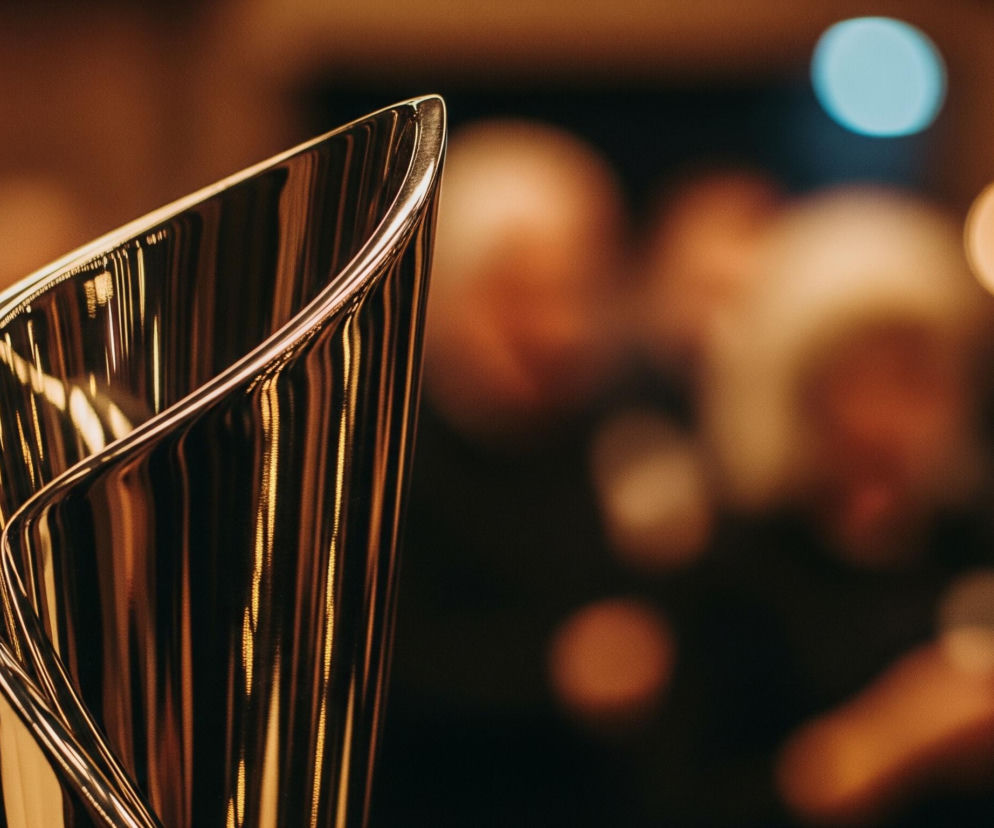Lewa strona kadru przedstawia fragment połyskującej, metalicznej rzeźby lub trofeum o zakrzywionych, opływowych kształtach, której powierzchnia odbija pionowe pasma światła w odcieniach złota i brązu. Element ten jest ostro zogniskowany, ukazując detale gładkiej, odbijającej światło krawędzi. Prawa strona obrazu to rozmyte tło, gdzie widać nieostre plamy świetlne w kolorach pomarańczowym, złotym oraz jedną wyraźną, jasnoniebieską plamę. Całość utrzymana jest w ciepłej kolorystyce, z dominacją brązów i pomarańczy.