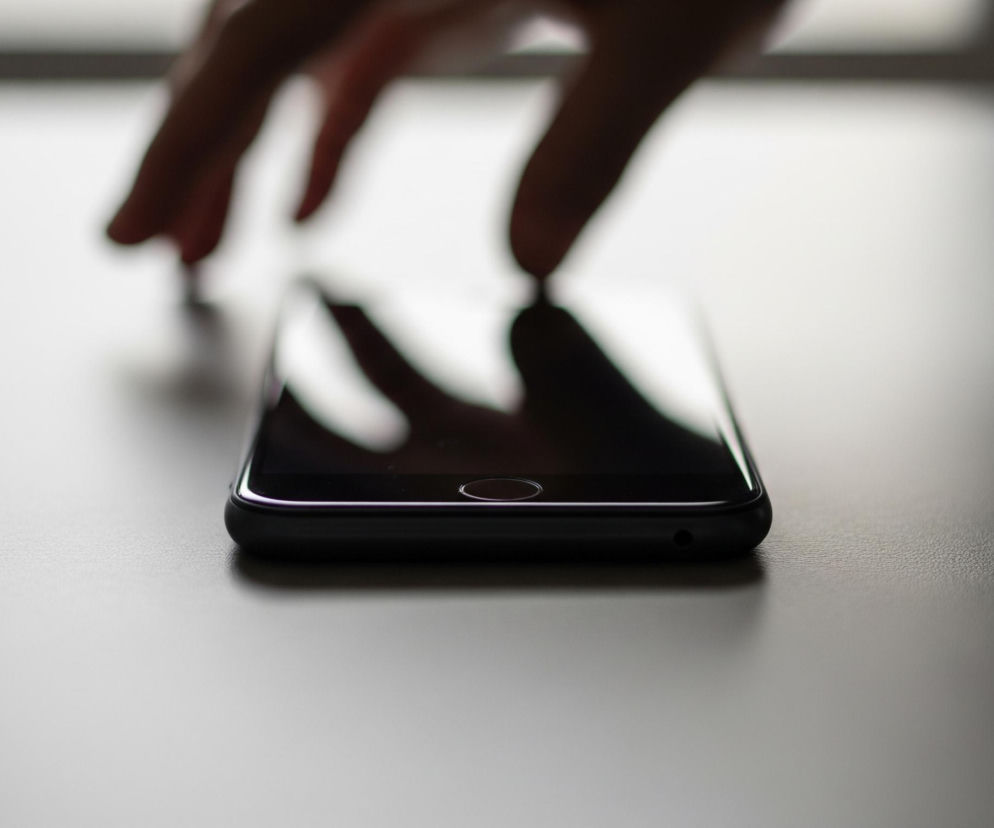 Ciemny smartfon leży płasko na jasnoszarym blacie, widoczny od boku z włączonym ekranem, który odbija światło. Górna krawędź urządzenia jest nieco ścięta. W tle widać rozmazaną dłoń z palcami rozciągniętymi nad telefonem, a także rozmyte tło z jasnym prześwitem w górnej części kadru. Blat jest częściowo oświetlony, tworząc łagodne przejście od jasnej szarości do ciemniejszego odcienia w prawym dolnym rogu.