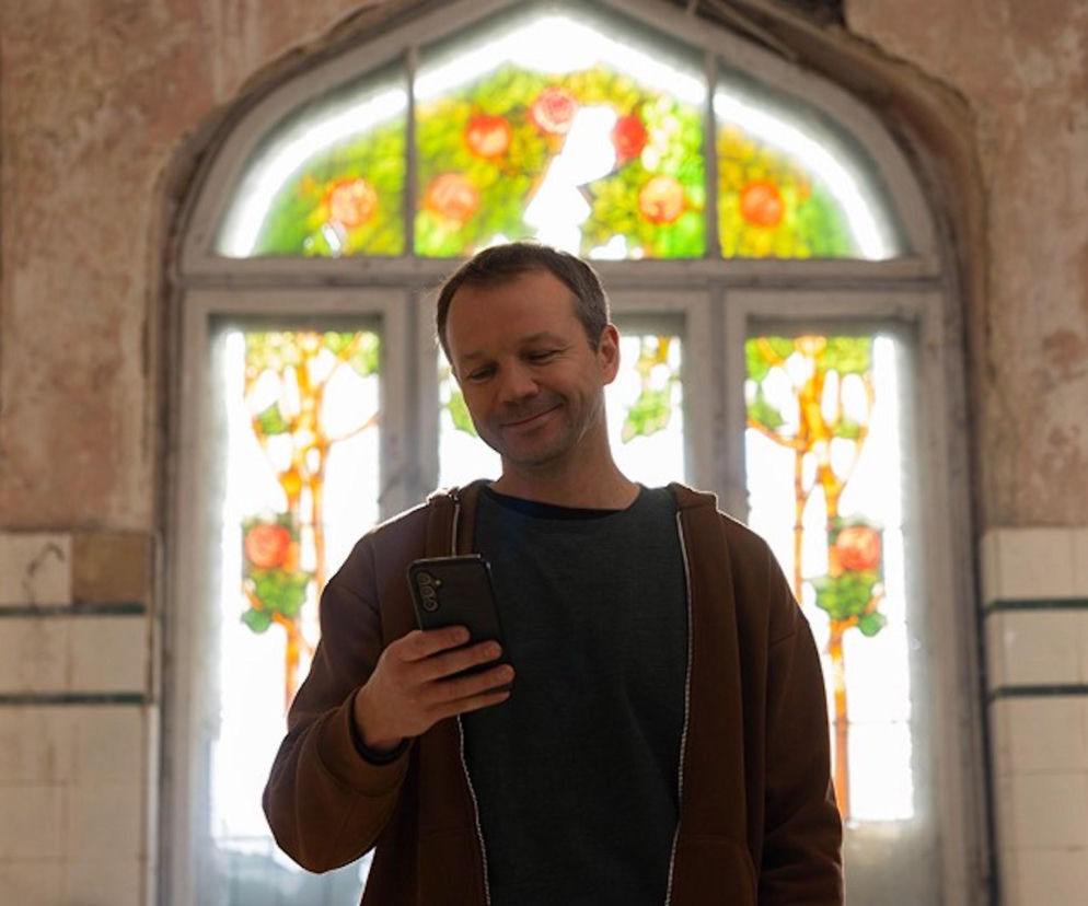 Uśmiechnięty Mateusz (Lesław Żurek) z serialu Pierwsza miłość, trzymający smartfon, na tle witrażowego okna w zrujnowanym wnętrzu. Scena sugeruje, że psychopata Mateusz wyśle mroczną wiadomość do Emilki, co można śledzić na stronie Super Seriale.