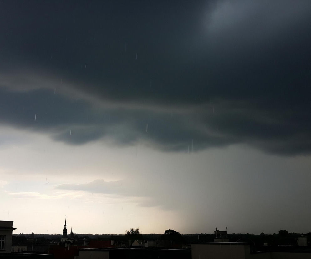 Deszcz pada z ciemnych, ołowianych chmur dominujących niebo, zajmując ponad połowę kadru u góry. Długie, cienkie krople deszczu są widoczne w powietrzu, zwłaszcza w górnej części obrazu. Poniżej rozciąga się miejski krajobraz z ciemnymi sylwetkami budynków i drzew, z wyraźnie widoczną wieżą kościoła po lewej stronie. Horyzont jaśnieje nieco, ukazując jaśniejsze niebo przechodzące w szarość.