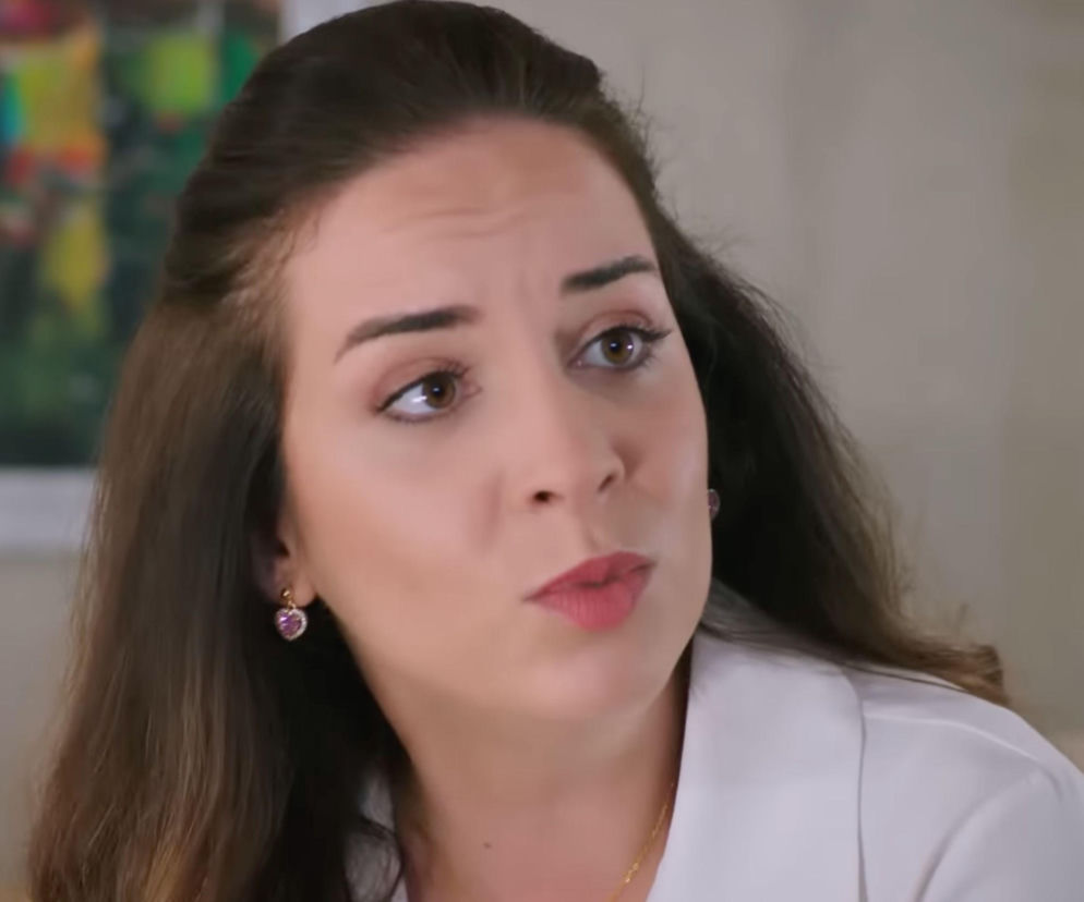 Kobieta z długimi, brązowymi włosami i kolczykami w kształcie serca, ubrana w białą bluzkę, patrzy w prawo z zamyślonym wyrazem twarzy. Odnosi się to do postaci Beyzy z serialu Panna Młoda, która w 79. odcinku przedstawia kontrowersyjny plan. Więcej o tym na Super Seriale.