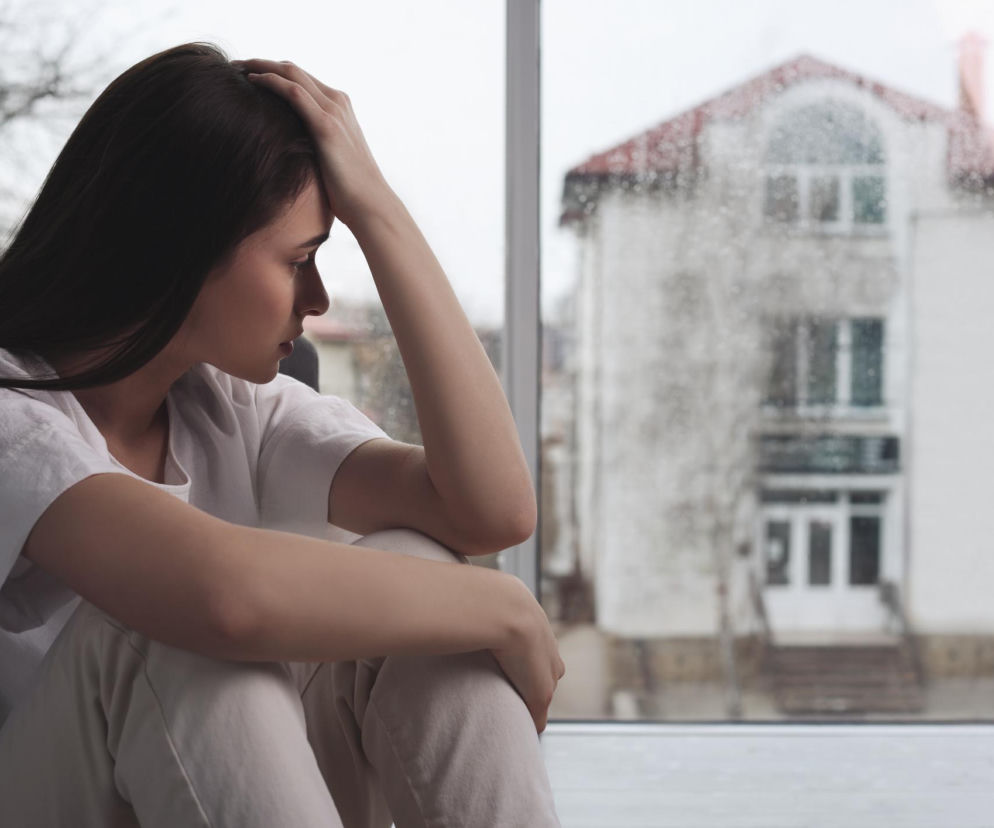 Zasmucona kobieta siedząca przy oknie podczas deszczu, dotykająca skroni, symbolizująca ból głowy i brak energii przed zmianą pogody. Dowiedz się, jak sobie radzić z meteoropatią, czytając artykuł na Poradnik Zdrowie.