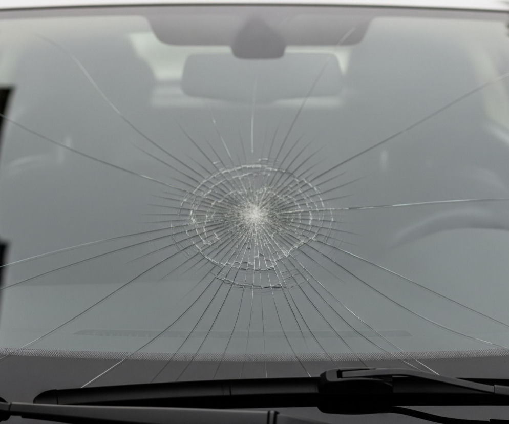 Pęknięta przednia szyba samochodu jest centralnym elementem obrazu, z uszkodzeniem w kształcie gwiazdy zlokalizowanym nieco powyżej jej środka. Od punktu uderzenia promieniują liczne, cienkie pęknięcia rozchodzące się na boki i w dół szyby. U dołu kadru widoczne są fragmenty czarnych wycieraczek samochodowych oraz ciemny pas u podstawy szyby, a w tle przez szkło można dostrzec niewyraźne, ciemne kształty wnętrza pojazdu.