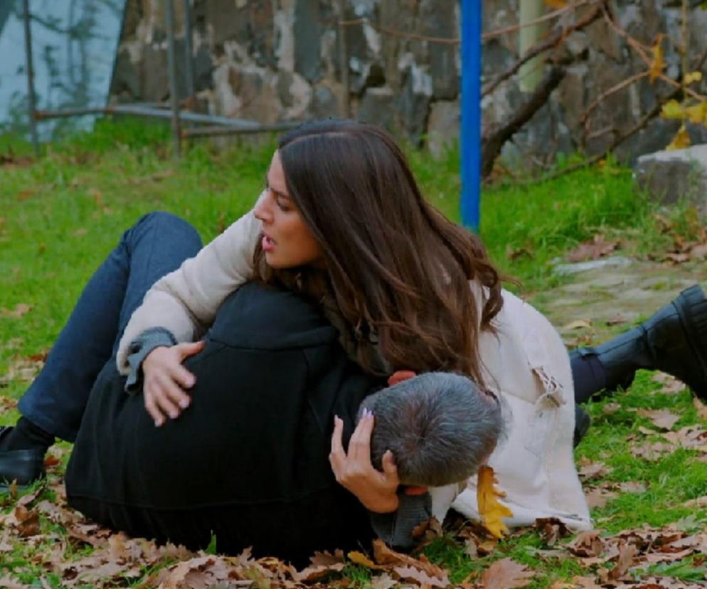 Zeynep rozpacza nad leżącym Bulentem po strzale Tahira. Kobieta z długimi, ciemnymi włosami obejmuje rannego mężczyznę, oboje leżą na trawie pokrytej jesiennymi liśćmi. Widoczna dramaturgia emocji w serialu Miłość i nadzieja na Super Seriale.