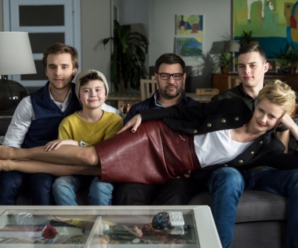 Uśmiechnięta Małgorzata Kożuchowska w czerwonej spódnicy, oparta na Tomaszu Karolaku w okularach, z młodymi Maciejem Musiałem, Adamem Zdrójkowskim i Mateuszem Pawłowskim na kanapie. To obsada serialu Rodzinka.pl, o której powrocie i nowościach przeczytasz na Super Seriale.