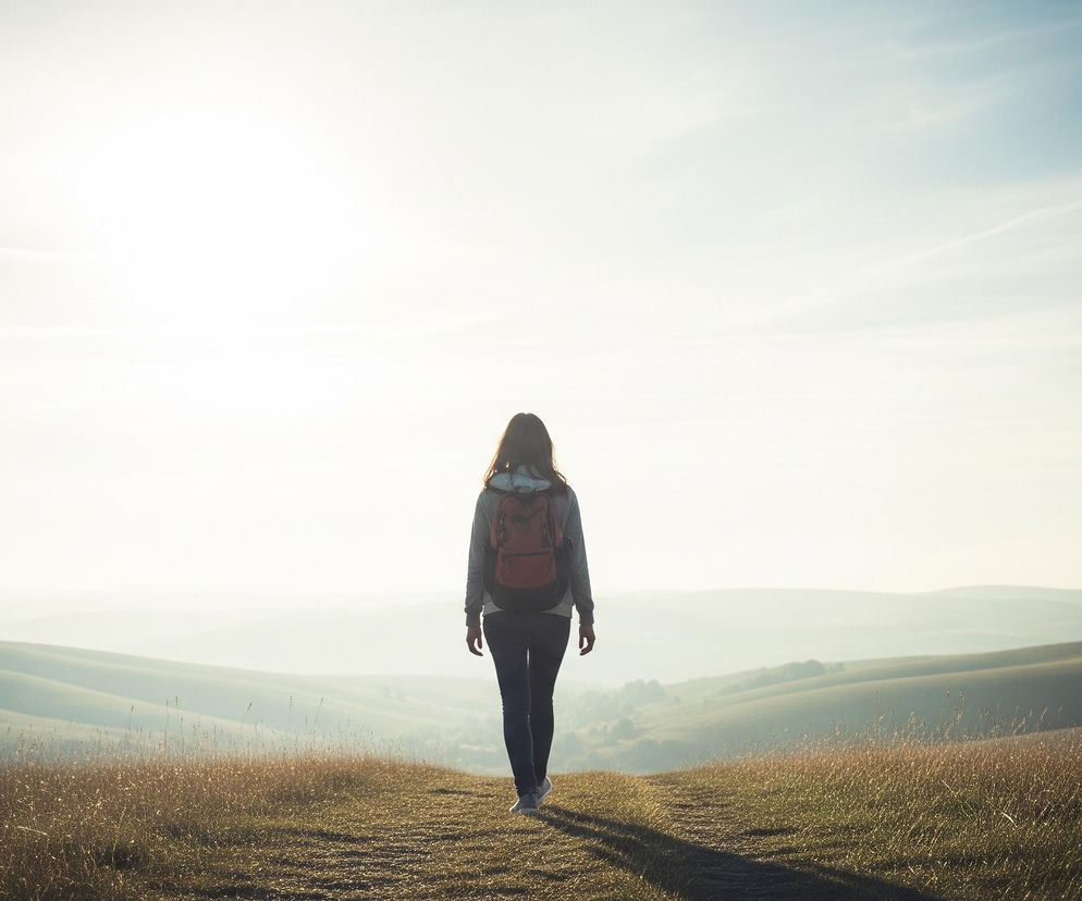 Kobieta z ciemnymi włosami, w szarej bluzie, ciemnych spodniach i z czerwonym plecakiem, idzie tyłem po polnej ścieżce, która prowadzi przez pagórkowaty krajobraz. Po obu stronach ścieżki rośnie sucha, żółto-brązowa trawa, oświetlona ciepłym, jasnym światłem, które dominuje w górnej części obrazu, rozjaśniając horyzont. W tle rozciągają się falujące wzgórza, stopniowo blednące w oddali, pod jasnym, częściowo błękitnym niebem z delikatnymi smugami chmur.