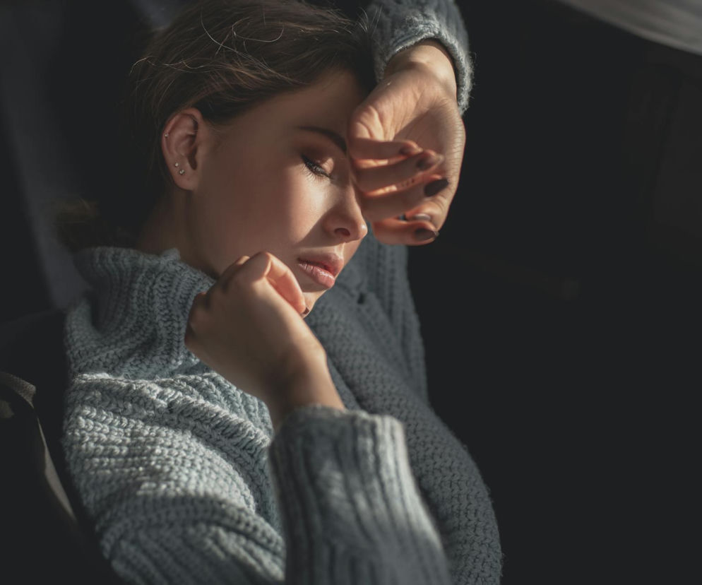 Kobieta w jasnoszarym swetrze z dłońmi zakrywającymi twarz, pogrążona w bólu migrenowym, co wskazuje na potrzebę ulgi od objawów. Dowiedz się, jak radzić sobie z atakami migreny bez tabletek, czytając artykuły na Poradnik Zdrowie.