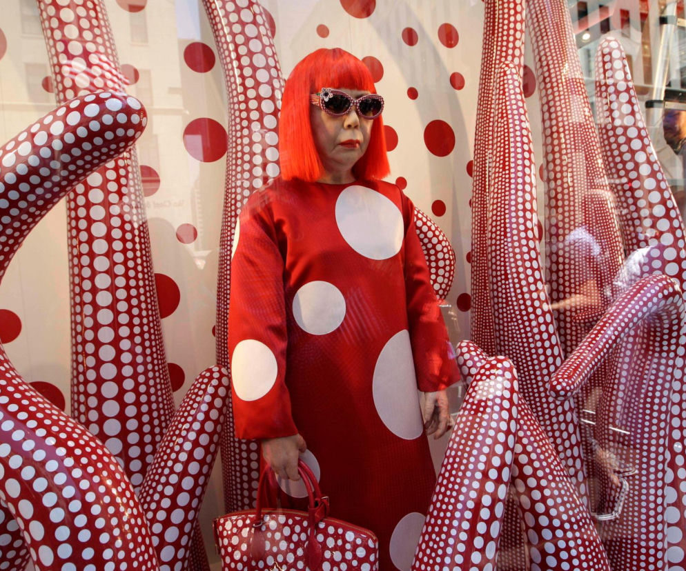 Artystka Yayoi Kusama w czerwonej peruce i sukience w kropki na tle swojej instalacji. O jej śmierci przeczytasz w Eska.