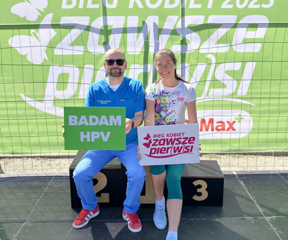Dr n. med. Wojciech Homola i Marzena Michalec pozują na tle zielonej banderoli z logo Biegu Kobiet Zawsze Pier(w)si. Trzymają transparenty promujące badania HPV, akcentując ważność profilaktyki raka szyjki macicy, o czym przeczytasz na Poradnik Zdrowie.