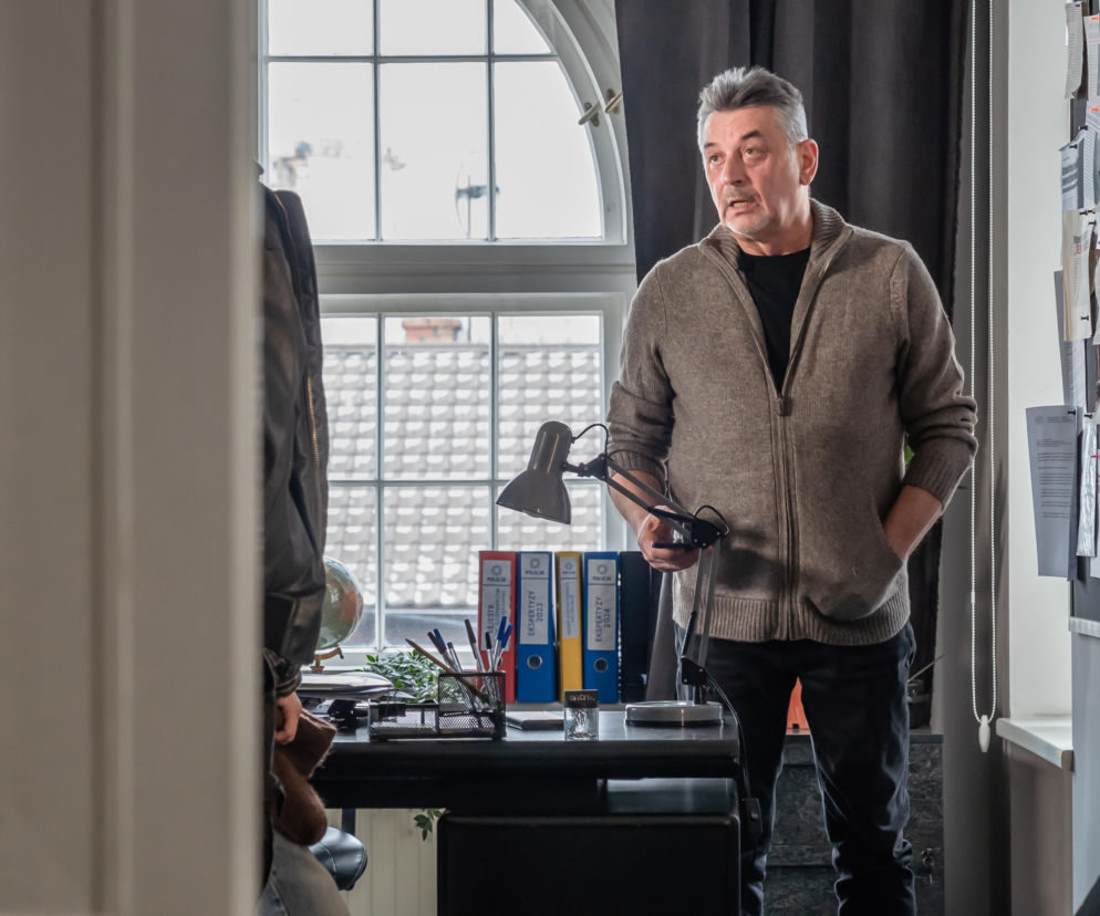 Mężczyzna w średnim wieku, w rozpiętym kardiganie i czarnym T-shircie, stoi w biurze obok biurka. Trzyma w ręku czarną lampkę biurkową, patrząc w bok z otwartymi ustami. W tle widoczne okno i segregatory z napisem EKSPERTYZY i POLICJA. Więcej informacji o serialach znajdziesz na Super Seriale.