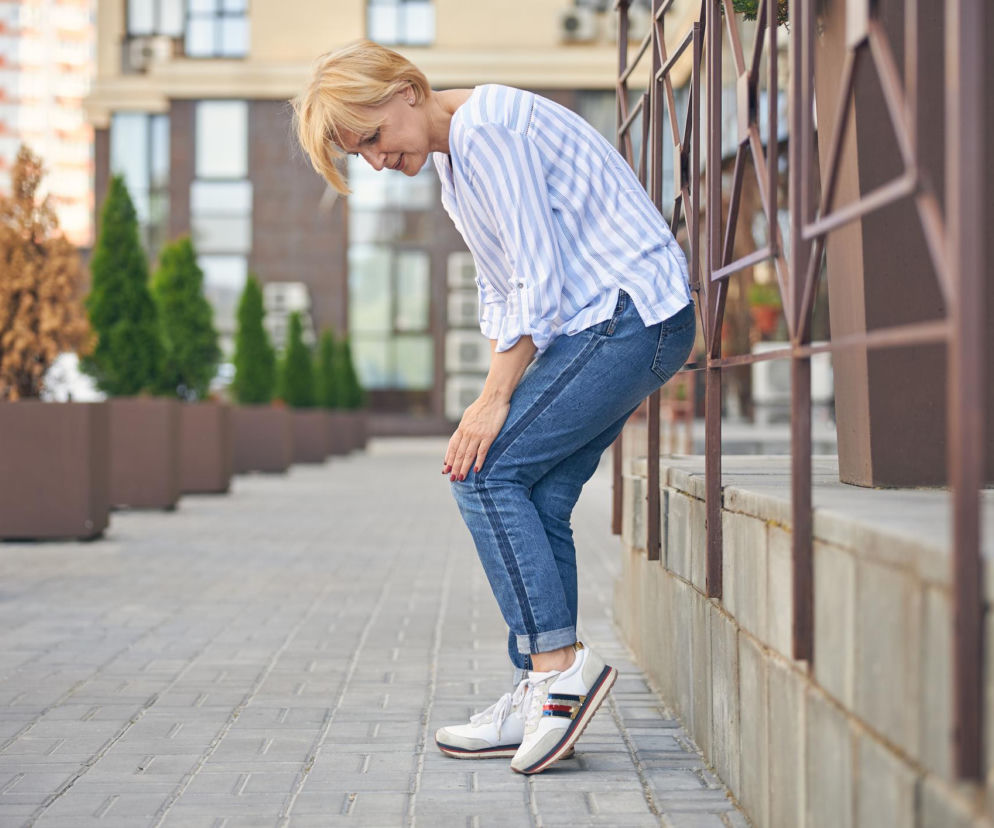 Kobieta z blond włosami w casualowym stroju, jeansach i koszuli, pochyla się, trzymając się za kolano, wyraźnie odczuwając ból podczas spaceru miejską ulicą. To symboliczny obraz syndromu oglądacza wystaw, o którym przeczytasz więcej na Poradnik Zdrowie.
