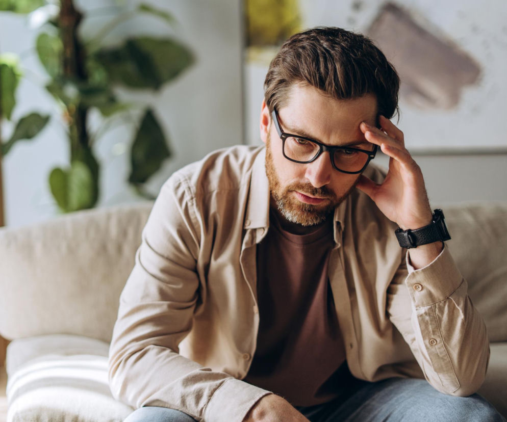 Mężczyzna w okularach z brodą, w beżowej koszuli i brązowej koszulce, siedzący na beżowej kanapie, z ręką na skroni, symbolizuje spadek libido i brak energii, najczęstsze objawy niskiego testosteronu, o czym przeczytasz na Poradnik Zdrowie.