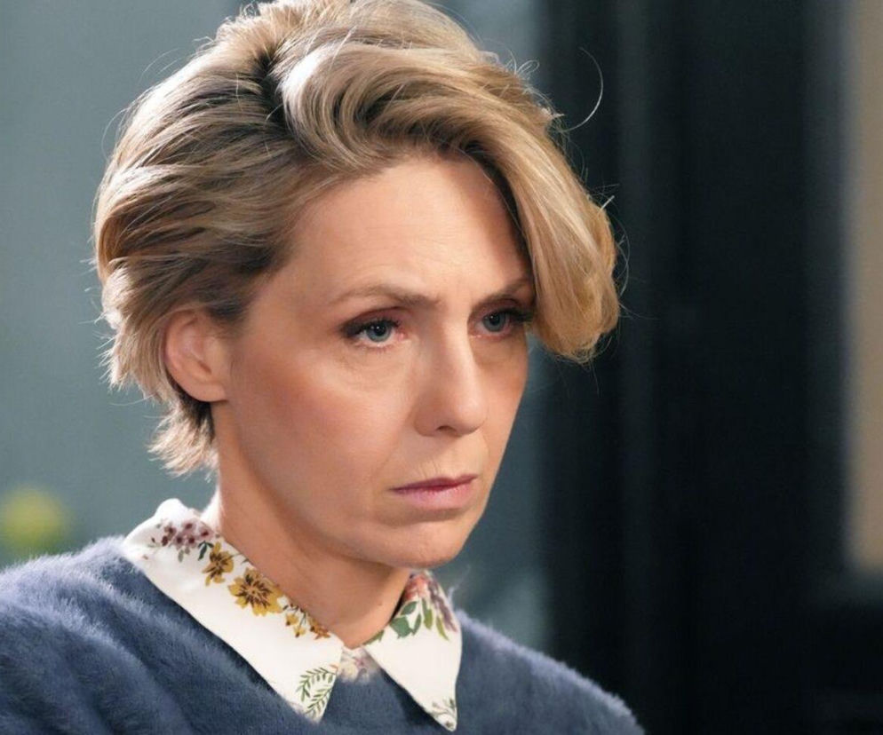 Blondwłosa Joanna Gleń jako Malwina w serialu „Barwy szczęścia”, z poważną miną, w niebieskim swetrze i białej koszuli w kolorowe kwiaty. Wygląda na zmartwioną po nieudanym zabiegu kosmetycznym, o czym przeczytasz więcej na portalu Super Seriale.