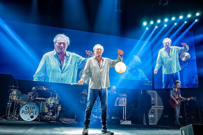 Ian Gillan, wokalista Deep Purple, na scenie podczas koncertu, trzymający gong i pałeczkę, ubrany w koszulę i jeansy. Za nim na ekranach widać jego powiększony wizerunek, a także pozostałych muzyków zespołu. O problemach ze wzrokiem wokalisty można przeczytać na portalu Poradnik Zdrowie.