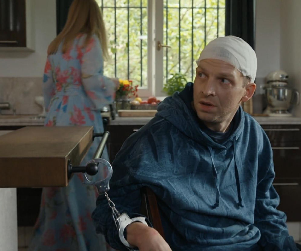 Michał Rolnicki jako Łukasz Sadowski z zabandażowaną głową i kajdankami na ręce, przykuty do blatu kuchennego. W tle kobieta w kwiecistej sukience. Scena z serialu Barwy szczęścia, której opis znajdziesz na Super Seriale.