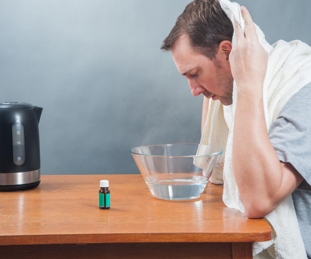 Mężczyzna wykonujący domową inhalację na zatkany nos, pochylony nad miską z gorącą wodą i przykryty ręcznikiem, z czarnym czajnikiem elektrycznym i buteleczką olejku eterycznego na drewnianym stole. Sprawdź, jak prawidłowo wykonać parówkę na Poradnik Zdrowie.