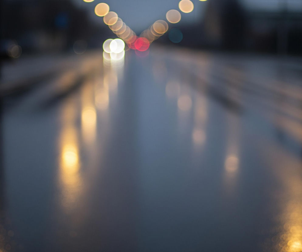 Rozmyty widok przedstawia oświetloną ulicę w deszczową noc lub o zmierzchu. Pośrodku kadru dominuje mokra, ciemna nawierzchnia drogi, odbijająca światła w postaci wydłużonych, rozmazanych smug i świetlistych plam. W górnej części obrazu widoczne są rozmyte punkty światła bokeh w kolorach żółtym, białym i czerwonym, biegnące łukiem od lewej do prawej strony, co sugeruje ruch pojazdów lub oświetlenie uliczne. Po prawej stronie, na horyzoncie, zarysowany jest niewyraźny kształt samochodu.