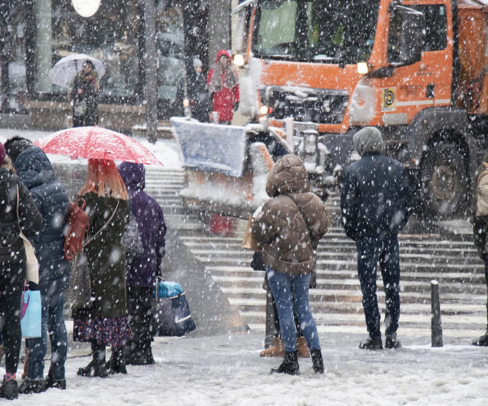 Grupa osób w ciepłych ubraniach, w tym kobieta z czerwoną parasolką, stoi na zaśnieżonym przejściu dla pieszych, podczas gdy w tle widać pług śnieżny. Scena ilustruje ekstremalne mrozy i trudne warunki pogodowe zapowiadane przez IMGW, o których można przeczytać na portalu Poradnik Zdrowie.
