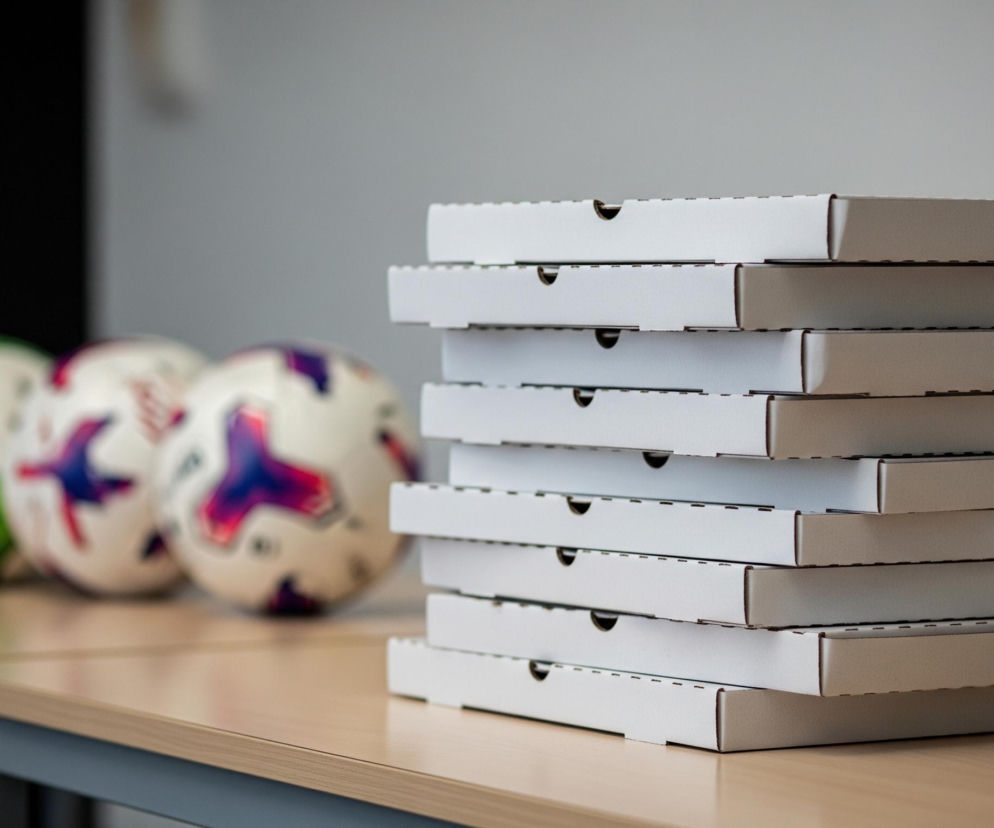 Stos białych, prostokątnych pudełek z tektury falistej, ustawionych pionowo, dominuje po prawej stronie kadru. Pudełka mają charakterystyczne wycięcia na krawędziach, służące do zamknięcia. Po lewej stronie, w tle i poza ostrością, widoczne są trzy piłki sportowe: jedna zielono-biała, a dwie pozostałe białe z fioletowymi i czerwonymi wzorami. Wszystkie obiekty spoczywają na jasnobrązowym, drewnianym blacie stołu, wspartym na ciemnych, metalowych nogach. Tło stanowi jasna, jednolita ściana, przechodząca w ciemniejszy obszar po lewej stronie, gdzie widoczny jest czarny lub bardzo ciemny element.