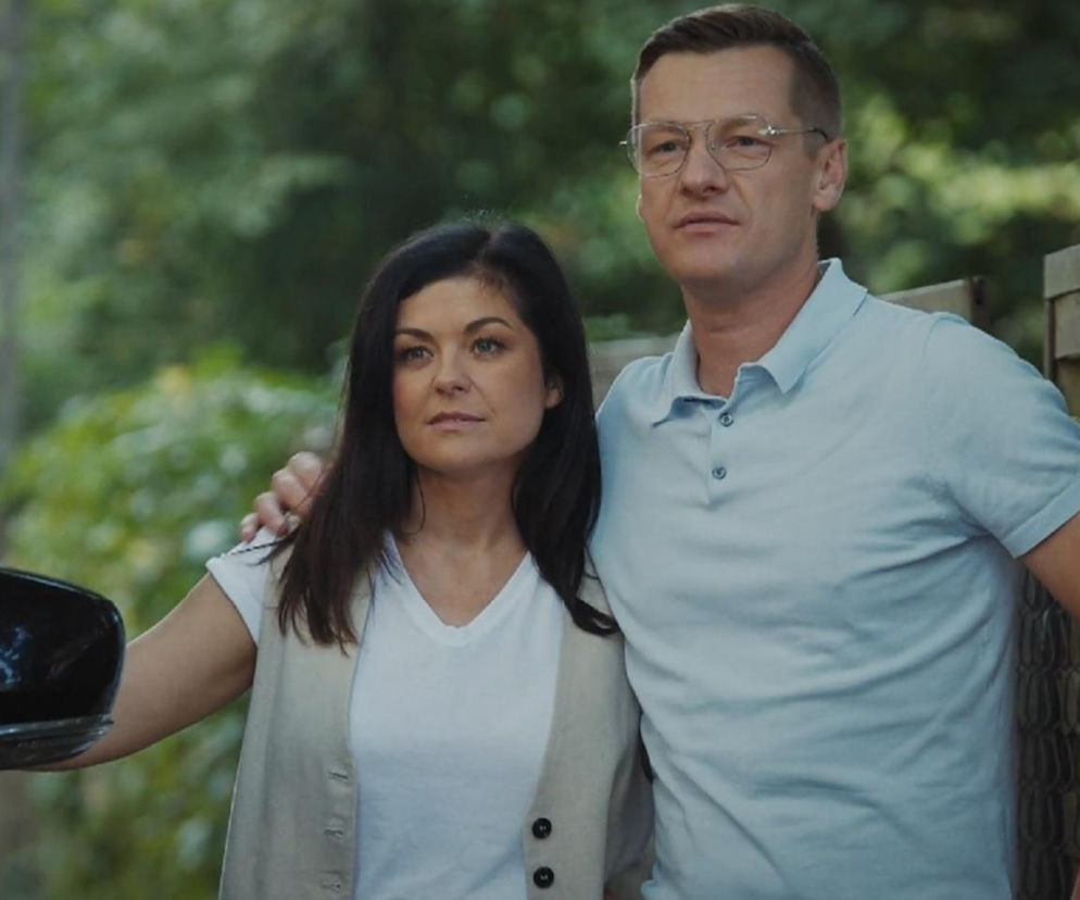 Kobieta z ciemnymi włosami w białym T-shircie i beżowej kamizelce, oparta o czarny samochód, oraz mężczyzna w okularach i błękitnej koszulce polo stojący obok, co zapowiada nowych sąsiadów Kingi i Piotrka w M jak miłość. Więcej informacji o serialu znajdziesz na stronie Super Seriale.