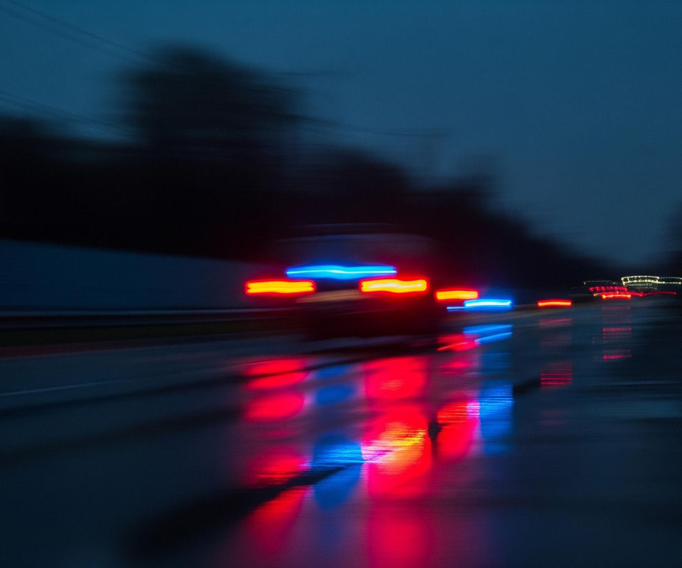 Rozmyte, dynamiczne ujęcie drogi w ciemności, z intensywnymi smugami czerwonych i niebieskich świateł odbijającymi się na mokrej nawierzchni. Główne źródło światła pochodzi od pojazdu znajdującego się w centrum kadru, którego rozmazane światła awaryjne są wyraźnie widoczne. Otoczenie drogi jest ciemne, z zamazanymi zarysami drzew i budynków po bokach, co podkreśla wrażenie szybkości i ruchu. Całość tonie w ciemnoniebieskich i granatowych barwach, z kontrastującymi jaskrawymi światłami.