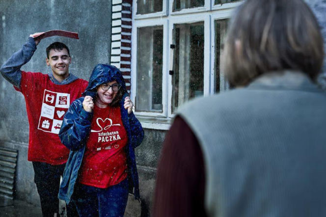 Dwoje uśmiechniętych wolontariuszy Szlachetnej Paczki odwiedza starszą osobę. O naborze przeczytasz na Radioplus.