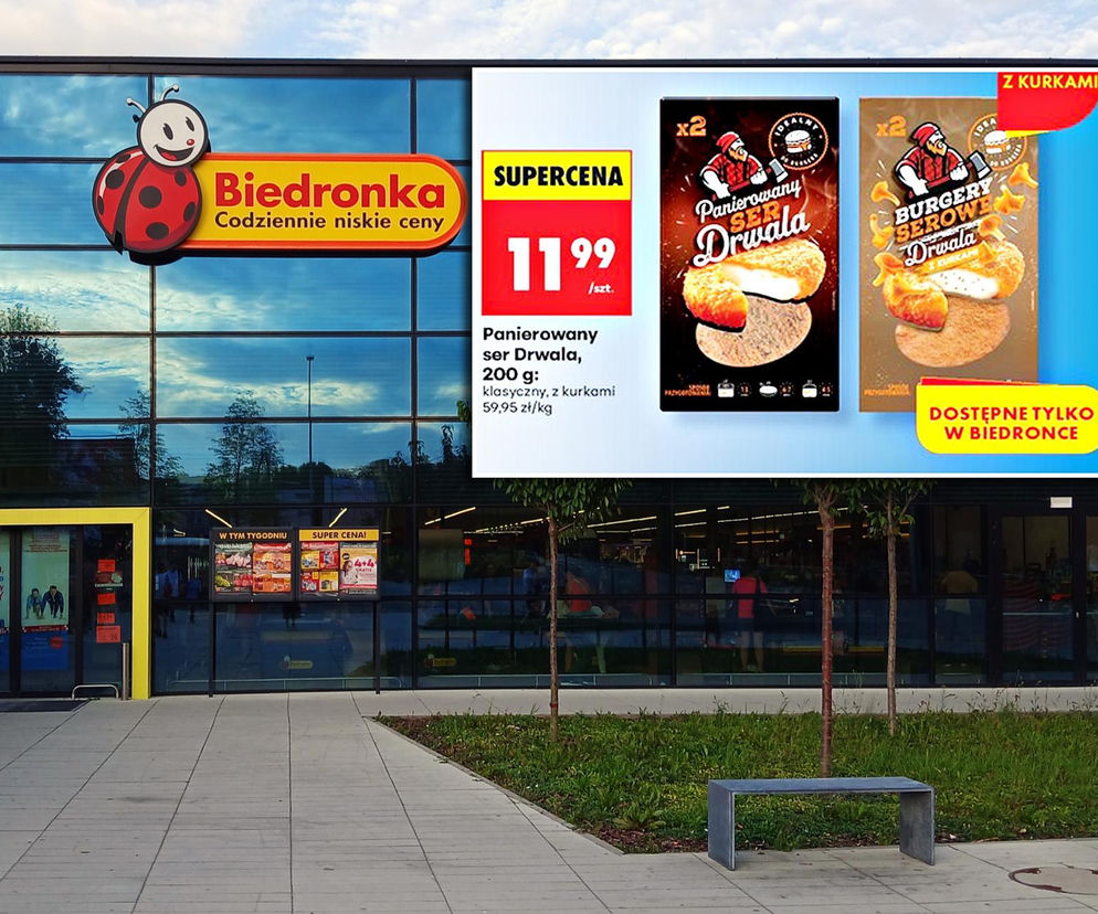 Elewacja sklepu Biedronka z dużym logo i szyldem Codziennie niskie ceny, a na niej reklama promocyjna burgerów serowych Drwala. Widać opakowania klasycznej wersji oraz z kurkami w Supercenie 11,99 zł za sztukę. Więcej o nowościach przeczytasz na Super Biznes.