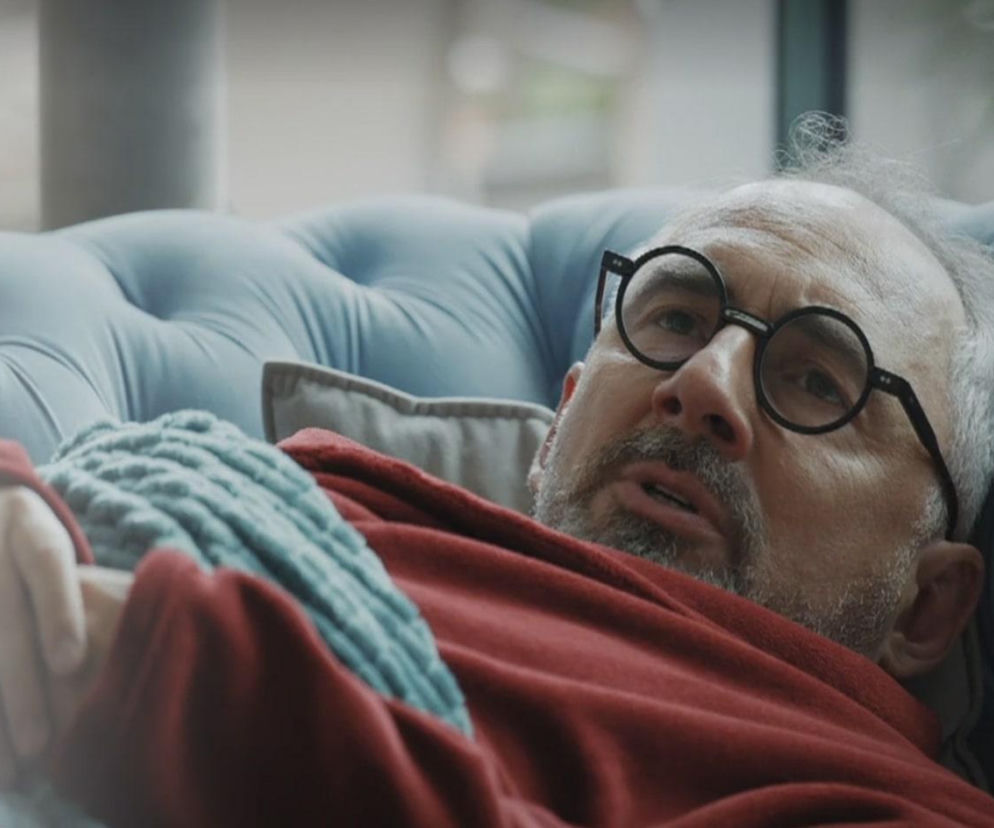 Aktor Jacek Kopczyński jako Adam Werner, leżący na niebieskiej kanapie w czerwonym szlafroku i pod niebieskim kocem, z wyrazem bólu na twarzy. Mężczyzna w okularach, z siwiejącymi włosami i brodą, patrzy w górę. Więcej o losach Wernera na Super Seriale.