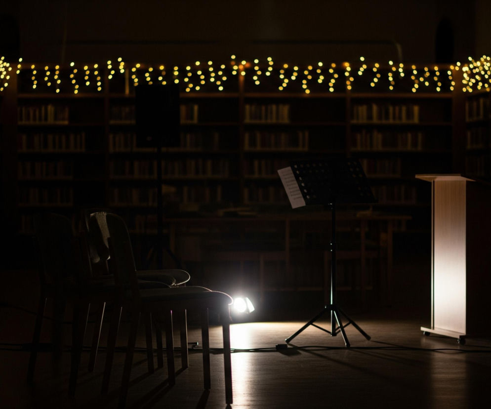 Scena przedstawia wnętrze ciemnej biblioteki nocą, oświetlone jedynie przez nieliczne, strategicznie rozmieszczone źródła światła. W centrum kadru widoczny jest pulpit na nuty z otwartym arkuszem muzycznym, stojący na trójnożnym statywie. Obok, po prawej stronie, znajduje się jasny, pionowy podest z drewna, oświetlony od wewnątrz lub z przodu, a za nim fragment krzesła z jasnoszarym obiciem. Na pierwszym planie, po lewej, ustawione są dwa ciemne krzesła, częściowo widoczne w kontrze, z których jedno jest wyraźnie oświetlone od tyłu przez punktowe światło z lampy stojącej na podłodze. W tle, wzdłuż ścian, ciągną się półki z książkami, które są niemal całkowicie pogrążone w cieniu, a nad nimi, po obu stronach kadru, rozciąga się girlanda z drobnych, ciepłych światełek, nadając wnętrzu świąteczny lub intymny charakter.