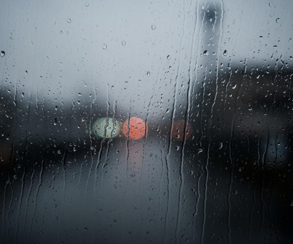 Przednia szyba samochodu jest pokryta licznymi kroplami deszczu i smugami wody, tworzącymi efekt rozmycia widoku. W tle, poprzez warstwę wody, widać niewyraźne, zamazane światła: dwa pomarańczowe i jedno zielone, sugerujące sygnalizację świetlną lub światła pojazdów. Ogólny kolor tła jest szary i ciemny, z delikatnymi odcieniami niebieskiego i czarnego, wskazując na pochmurny dzień lub zmierzch.
