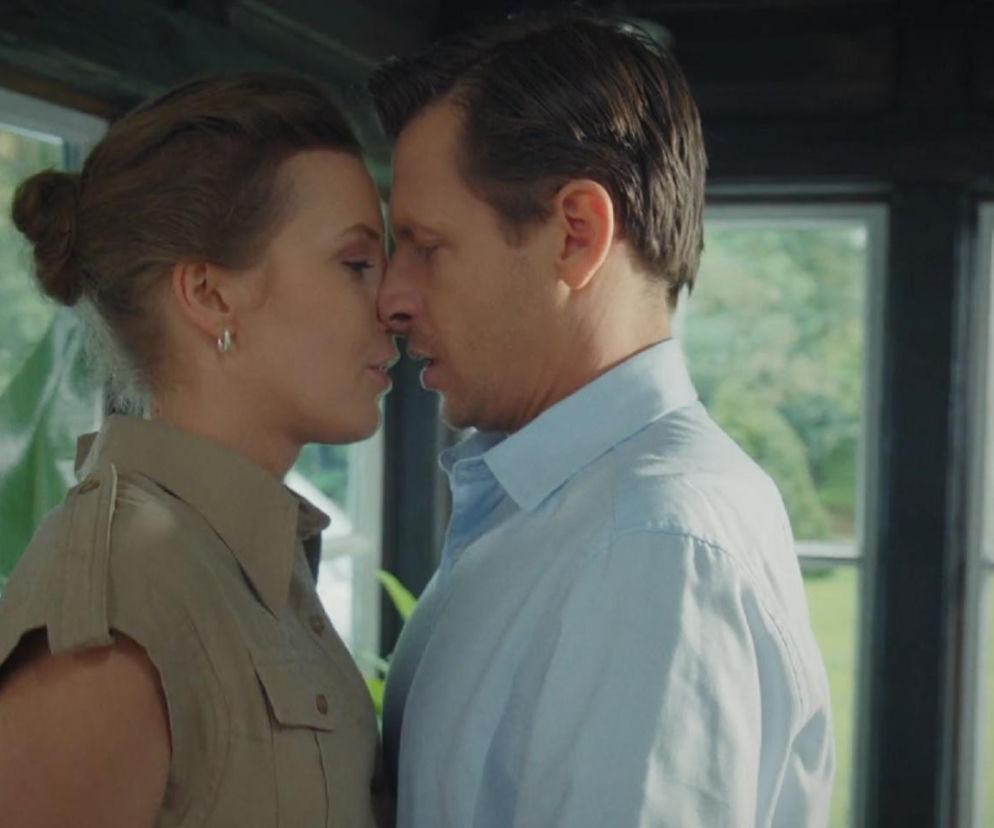 Bliskie zbliżenie Natalii Mostowiak i Adama Karskiego z serialu M jak miłość, gotowych na pocałunek, z zielenią za oknem w tle. Scena romansu i ukrywanego związku, który wyjdzie na jaw w 1920 odcinku, dostępnym na portalu Super Seriale.