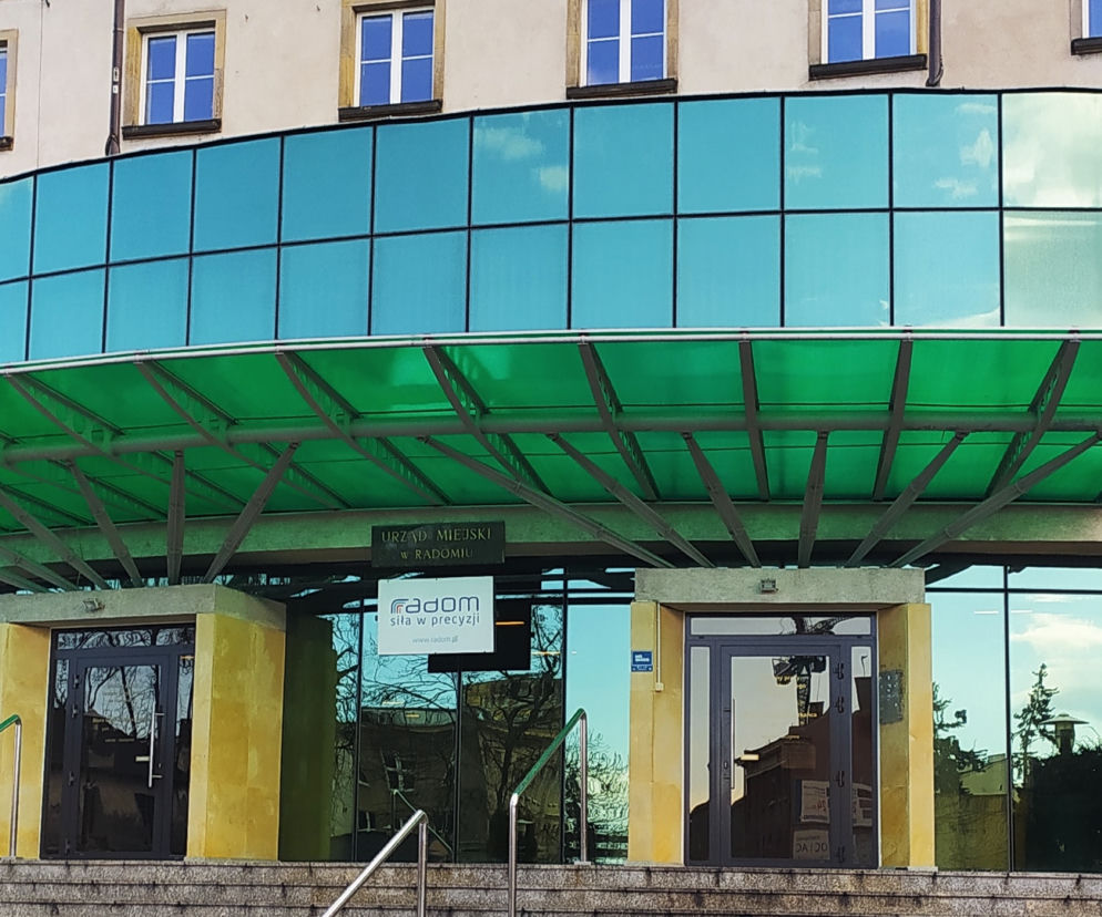 Fasada Urzędu Miejskiego w Radomiu z zielonym zadaszeniem wejścia i błękitnym szkłem, u góry beżowa ściana z oknami. Na tabliczce logo Radom siła w precyzji. Więcej informacji o udzieleniu wotum zaufania przeczytasz na naszym portalu.