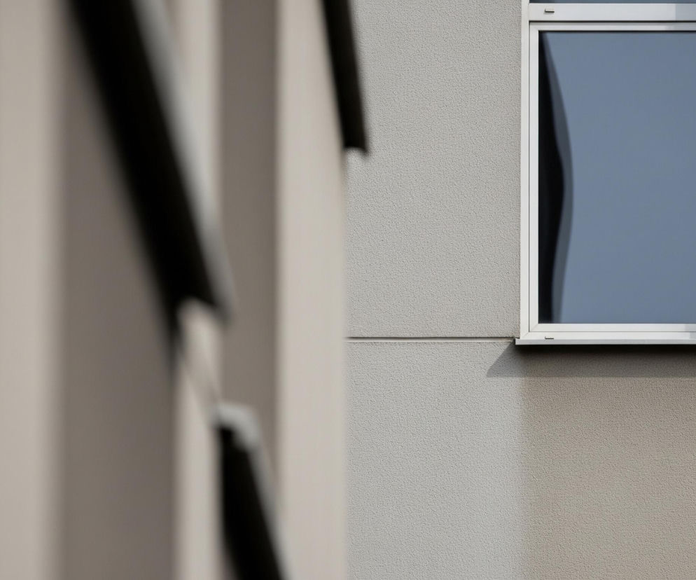 Na zdjęciu widoczne jest pojedyncze, prostokątne okno z szarą ramą i niebieskim szkłem, umiejscowione na jasnoszarej, ziarnistej ścianie po prawej stronie kadru. Po lewej stronie, poza ostrością, znajdują się pionowe, beżowe i ciemne kształty, sugerujące fragmenty innej struktury lub budynku. Od okna rzuca się ciemny cień na ścianę poniżej, a odbicie w szybie pokazuje falistą, ciemniejszą formę.