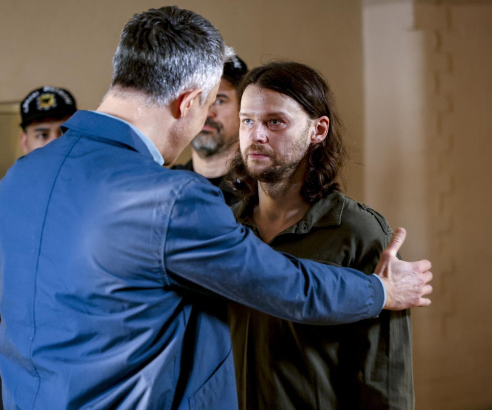 Mateusz Kościukiewicz jako Paweł Krzyżanowski w serialu Pierwsza miłość, stoi w areszcie policyjnym, a mężczyzna w niebieskiej kurtce trzyma go za ramię. Za nimi widać postacie w mundurach. Kontekst odcinka Pierwszej miłości znajdziesz na Super Seriale.