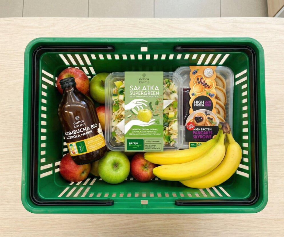 Zielony koszyk na zakupy wypełniony zdrowymi produktami: kombucha BIO, sałatka Supergreen, proteinowe pancakes oraz świeże jabłka i banany, odzwierciedlający zbilansowaną ofertę Żabki. Więcej o nowościach w Super Biznes.