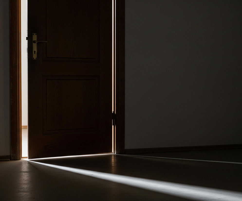 Ciemne wnętrze pomieszczenia z częściowo otwartymi, drewnianymi drzwiami w centrum kadru, skąd wpada jasne światło, tworząc smugę na podłodze. Lewa strona ściany w pokoju jest oświetlona, ujawniając jasny włącznik światła. Prawa strona kadru pozostaje w głębokim cieniu.
