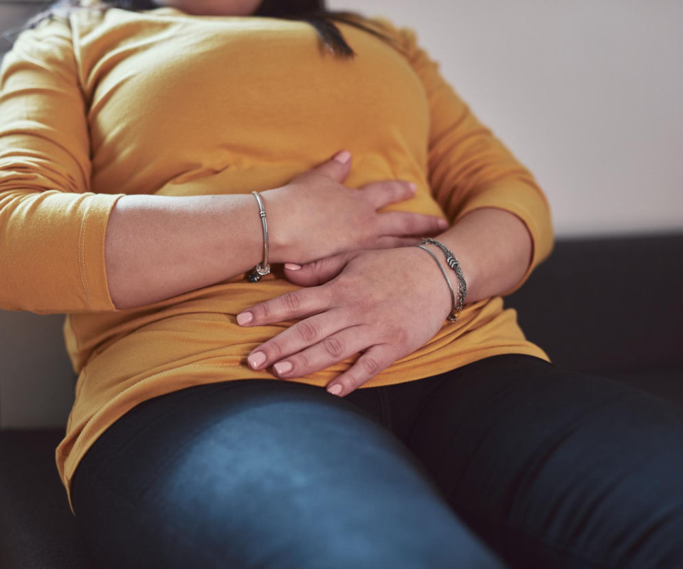 Kobieta w żółtej bluzce i ciemnych spodniach leży, trzymając ręce na brzuchu w geście bólu lub dyskomfortu. Zdjęcie ilustruje problemy z układem pokarmowym, takie jak wzdęcia czy ból brzucha. Na portalu Poradnik Zdrowie znajdziesz więcej informacji o objawach i przyczynach chorób trzustki.