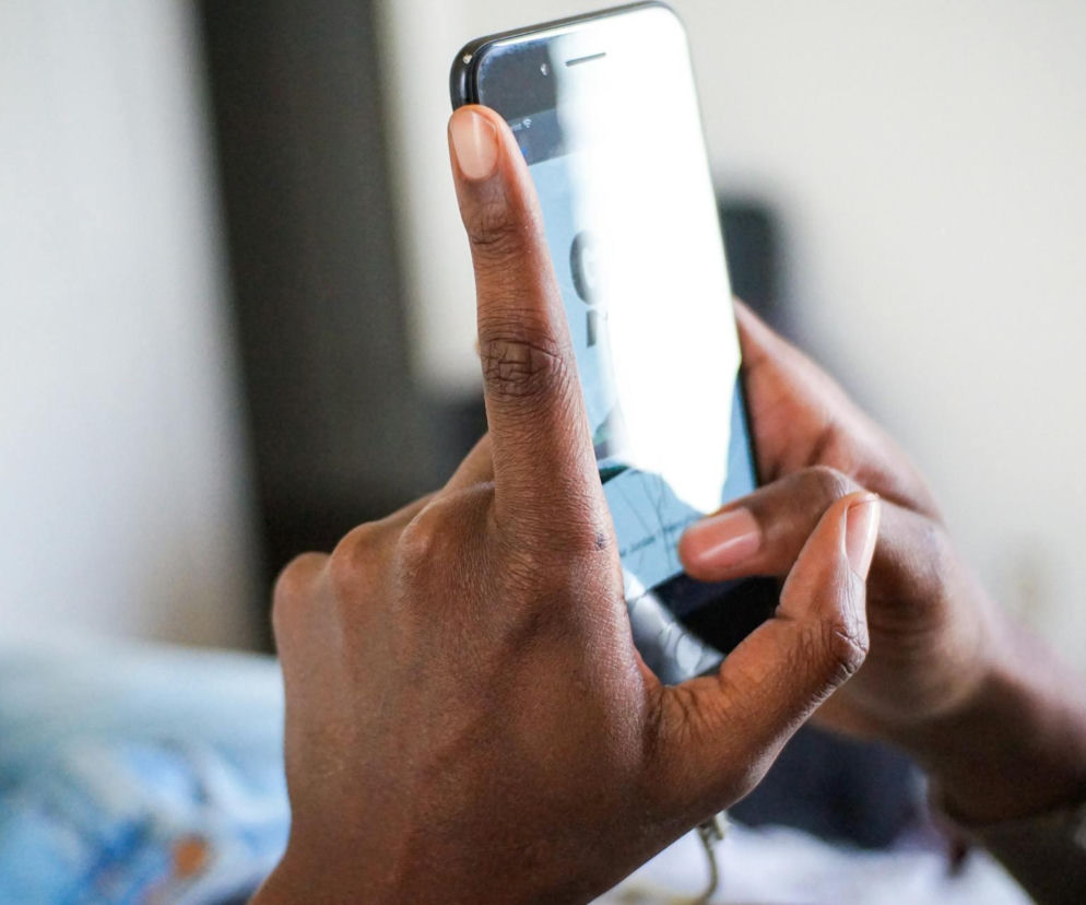 Ciemnoskóre dłonie trzymają smartfon i przewijają jego ekran. To częsty nawyk, który, jak wskazuje Poradnik Zdrowie, może prowadzić do spadku nastroju i frustracji z powodu ciągłego porównywania się z innymi.