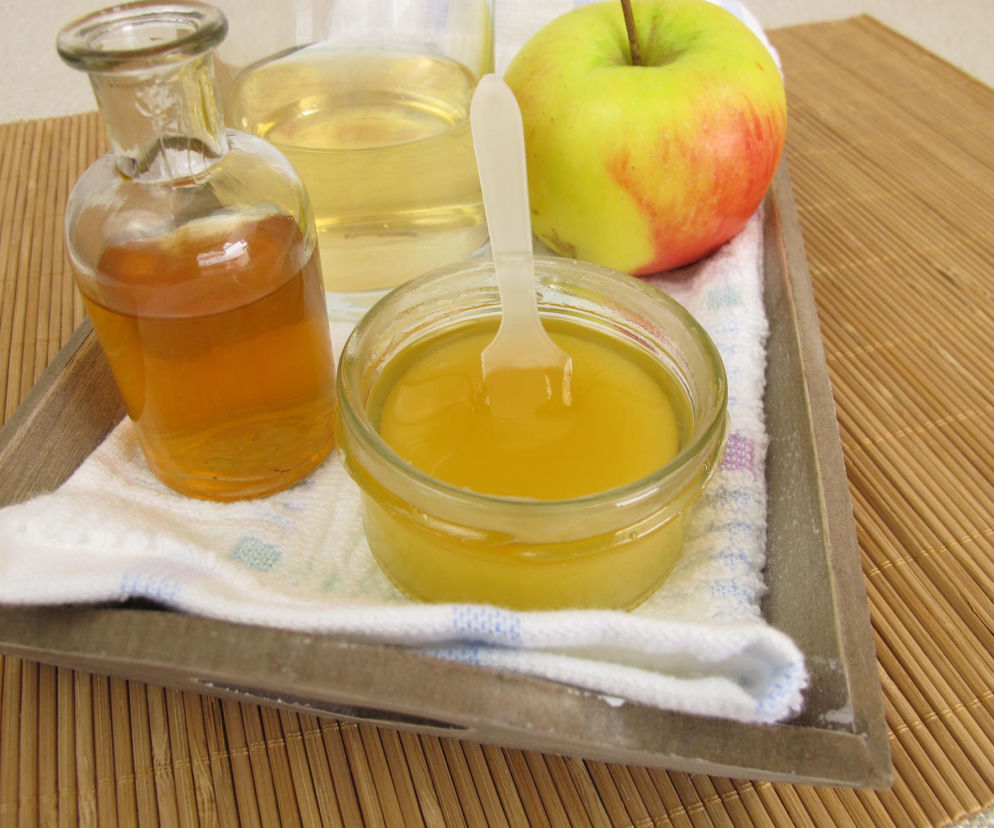 Ocet jabłkowy w butelce, miód w słoiczku z łyżeczką i świeże jabłko obok, ustawione na białej serwetce i drewnianej tacy, co symbolizuje naturalne składniki eliksiru zdrowia, o którym przeczytasz na Poradnik Zdrowie.