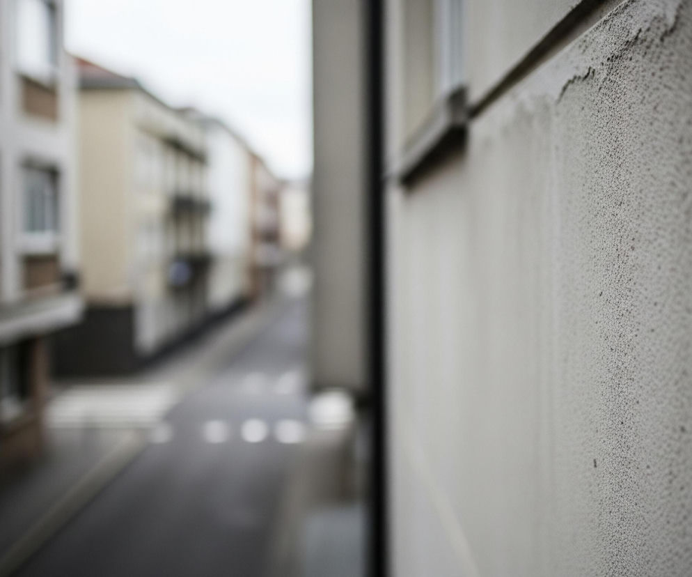 Prawa strona zdjęcia przedstawia fragment ściany budynku w jasnym odcieniu szarości z widoczną teksturą chropowatego tynku. Od góry widoczne jest zarysowanie ściany, a jej górna krawędź jest lekko ciemniejsza i wyraźniejsza. Po lewej stronie, w oddali, rozciąga się ulica otoczona kilkupiętrowymi budynkami o jasnych fasadach, z widocznymi oknami i balkonami. Na ulicy widoczne są białe linie i koła, prawdopodobnie oznaczenia poziome. Całość ujęcia jest lekko rozmyta poza pierwszym planem ściany.