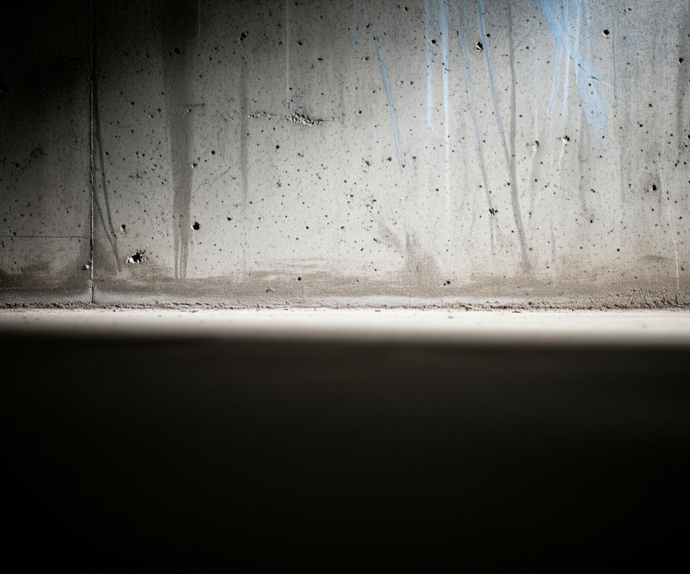 Szarobrązowa, betonowa ściana pokryta jest licznymi, nieregularnymi czarnymi plamkami i drobnymi dziurami. W górnej części ściany widoczne są pionowe smugi w odcieniach błękitu i bieli, niektóre z nich są cieńsze i zanikają ku dołowi, inne są grubsze i bardziej wyraźne. Linia styku ściany z podłożem jest nieregularna i usiana drobnymi fragmentami betonu i pyłu. Dolna połowa obrazu jest niemal całkowicie czarna, z delikatnym przejściem tonalnym od jasnoszarego paska na horyzoncie do głębokiego cienia.