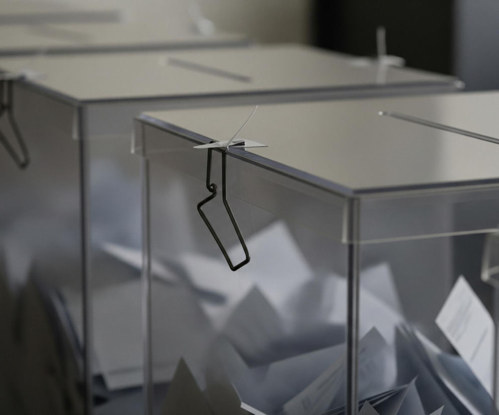 W rzędzie stoją cztery przezroczyste urny, częściowo wypełnione pogniecionymi białymi kartkami. Urny są wykonane z bezbarwnego tworzywa, z prostokątnymi otworami w górnej części każdej z nich. Na przodzie każdej urny, przy górnej krawędzi, widoczne są metalowe zapinki z drucianymi pętlami, służące do zamknięcia. Oświetlenie z góry tworzy odbicia na płaskich powierzchniach urn i cienie wewnątrz, a tło jest rozmyte.