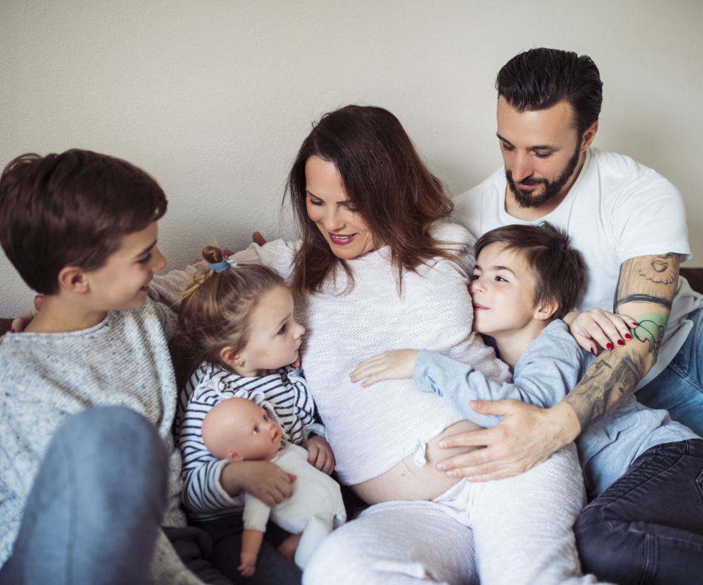 Szczęśliwa, wielodzietna rodzina z kobietą w ciąży na kanapie. Dwoje starszych dzieci i ojciec tulą się do brzucha matki. Artykuł o Karcie Dużej Rodziny, z której nowościami można zapoznać się na Super Biznes.