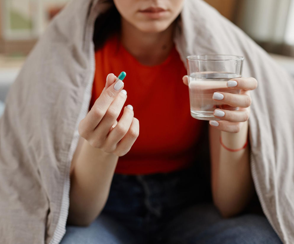 Kobieta w pomarańczowej koszulce, okryta kocem, trzymająca w dłoni zieloną kapsułkę i szklankę wody. Obraz ilustruje problem z połykaniem tabletek, o którym przeczytasz na portalu Poradnik Zdrowie.